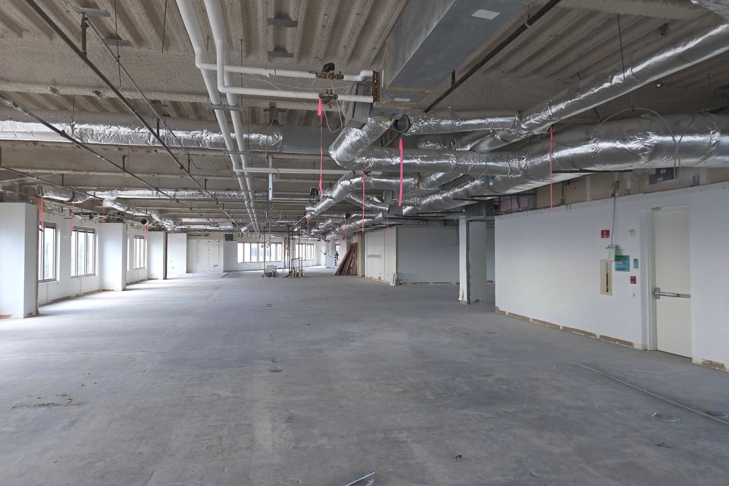 Empty commercial tenant-improvement floor with exposed insulated ductwork and a red fire alarm pull station and strobe on the far wall